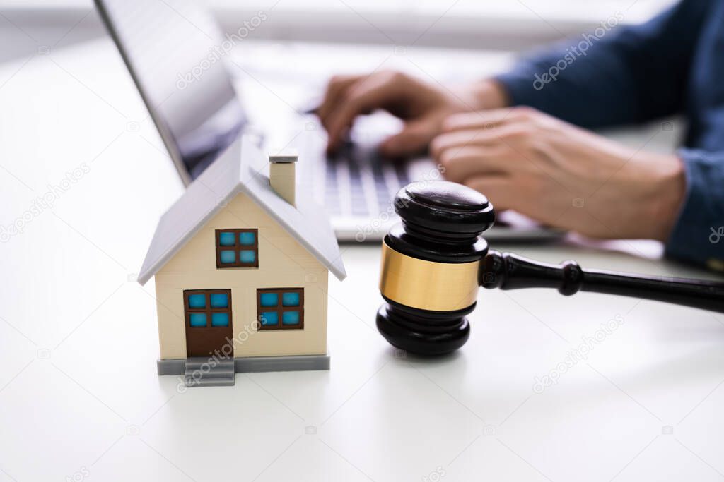 House Model With Gavel In Front Of A Businessperson Using Laptop On Wooden Desk