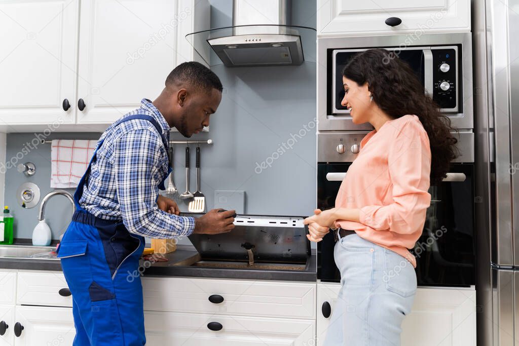 Woman Looking At Serviceman In Uniform Fixing Induction Stove In The Kitchen