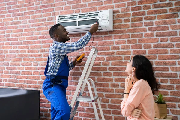 年轻女子看着男性技术人员修理安装在砖墙上的空调 — 图库照片