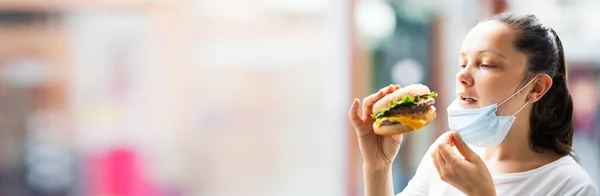 女性はレストランで顔マスクでファーストフードバーガーを食べる — ストック写真