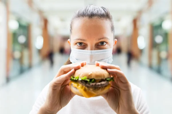 女性はレストランで顔マスクでファーストフードバーガーを食べる — ストック写真