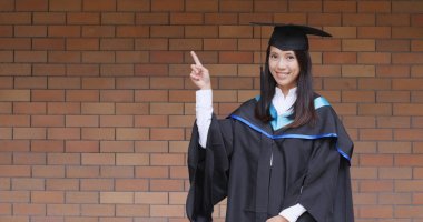 Mezuniyet elbisesi ile kırmızı tuğla duvarın üzerinden noktası gösterilen kadın