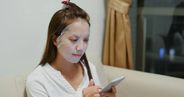 Vrouw toepassen gezichtsmasker en gebruik van mobiele telefoon thuis — Stockfoto