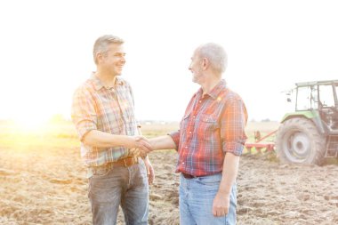 Çiftçiler çiftlikte el sıkışarak birbirlerini selamlıyorlar ve arka planda sarı mercek parlıyor. 