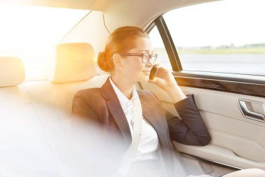 Genç çekici bir iş kadını, arka planda sarı bir mercekle arabada otururken akıllı telefondan konuşuyor. 