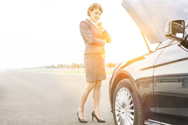 Mature businesswoman looking at breakdown car engine on road against sky