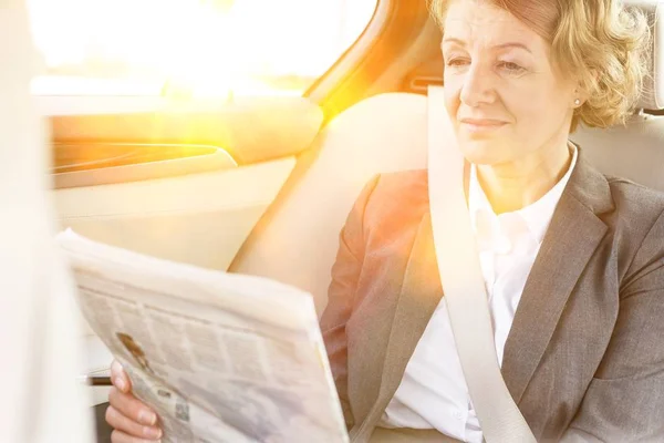 Mature businesswoman reading newspaper while sitting on car with yellow lens flare in background