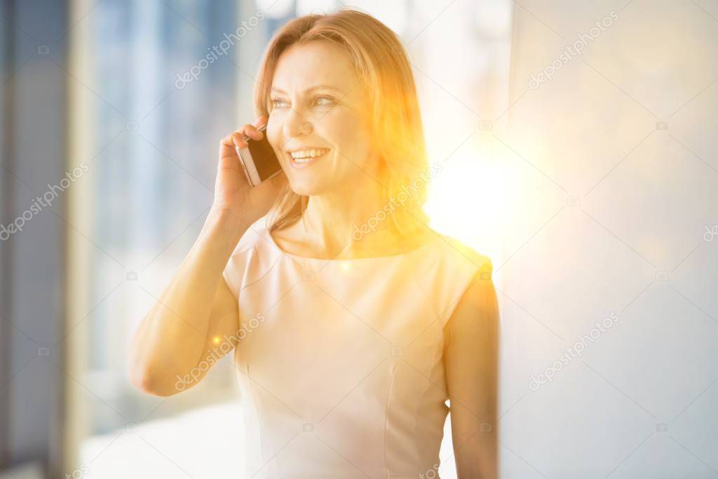 Businesswoman smiling while talking on smartphone in office with yellow lens flare in background