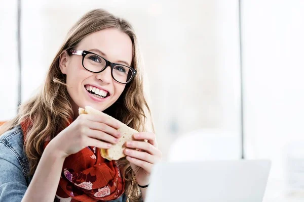 若いです魅力的なビジネス女性笑顔ながら食べるサンドイッチオン彼女の机でオフィス — ストック写真