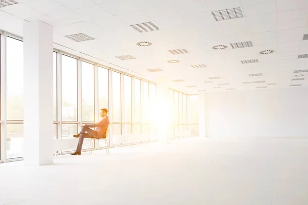 新しい空のオフィスの窓を見ながら椅子に座っている思慮深い若いビジネスマンの完全な長さ — ストック写真