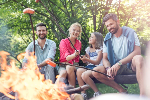 Happy family with male friend roasting sausages over campfire at park