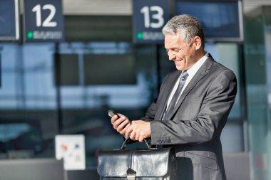 Olgun iş adamı havaalanında akıllı telefon kullanırken gülümsüyor.
