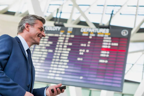 空港のフライトディスプレイ画面に立ちながらスマートフォンを使用して成熟した魅力的なビジネスマン — ストック写真