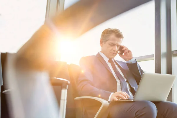 空港に着く前に彼のラップトップで座って作業している間にスマートフォンで話している成熟した魅力的なビジネスマン — ストック写真