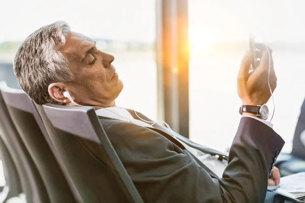 空港で搭乗するのを待っている間 彼のスマートフォンで音楽を聴く成熟したビジネスマンの肖像画 — ストック写真