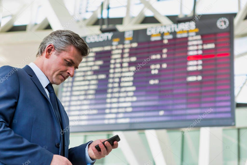 Mature attractive businessman using smartphone while standing against flight display screen in airport