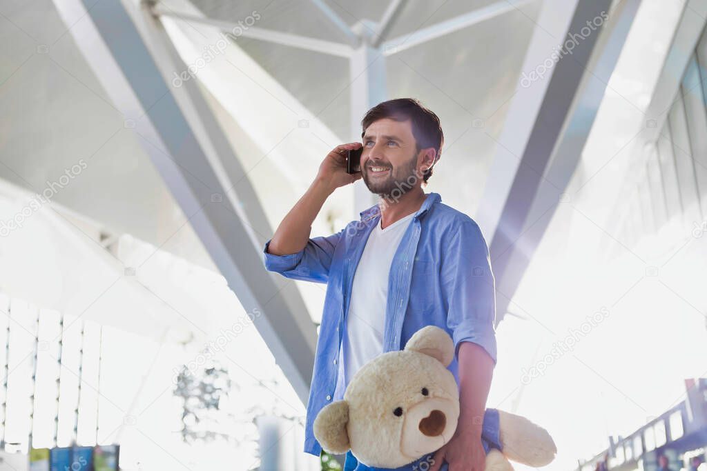 Portrait of man talking on smartphone while holding a teddy bear present for his daughter in airport
