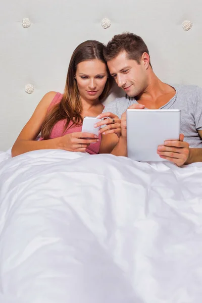 一个年轻迷人的女人在他的男朋友在床上玩数字平板电脑游戏时拿智能手机的画像 — 图库照片