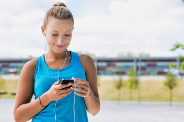Parkta akıllı telefonuyla müzik dinlerken ayakta duran çekici bir kadının portresi.