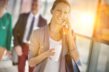 Genç çekici bir iş kadınının portresi. Akıllı telefondan konuşan ve meslektaşına karşı yürürken elinde bir fincan kahve tutan.