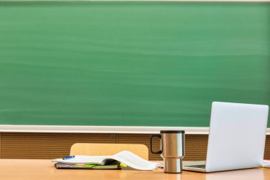 Profesör masasındaki bilgisayarın fotoğrafı. Taklacı ve kitapla sınıfta siyah tahtaya karşı.