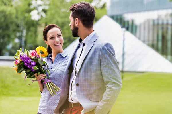 若い魅力的なビジネスカップルが公園を歩いている間 女性は花束を持っています — ストック写真