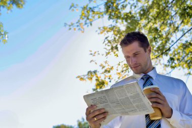 Mola sırasında parkta gazete okurken elinde bir fincan kahve tutan yakışıklı iş adamının portresi.