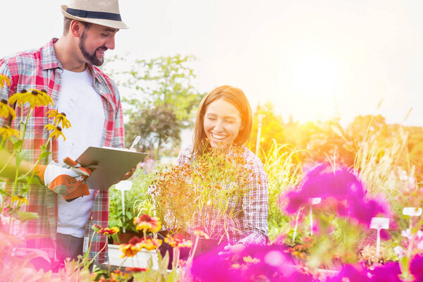 Portrait of mature gardener writing report on clipboard while female gardener arranging flowers on pot