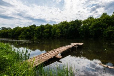 Yaz manzarası, bulutlar gökyüzünde yavaşça süzülüyor, bir nehir ve küçük bir iskele, yeşil ağaçlar.