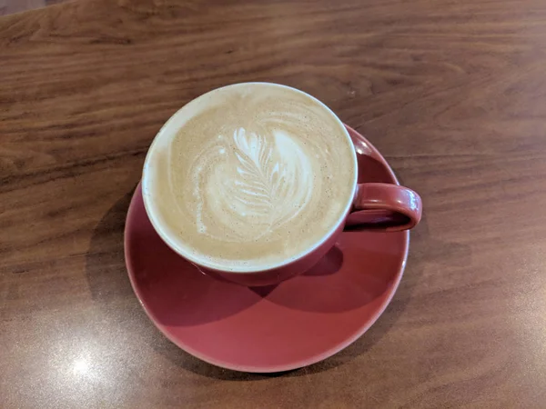 Red cup of Cappuccino on saucer with a leaf pattern in foam — Stock Photo, Image