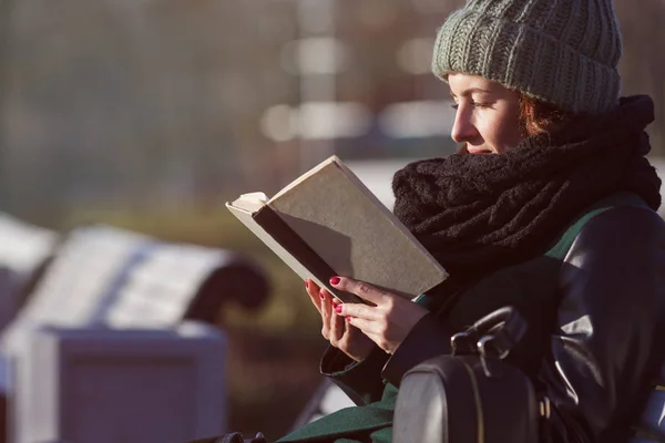 En flicka i en hatt sitter på en parkbänk och läser en bok. — Stockfoto