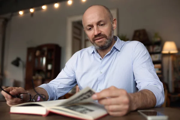 Um homem brutal com barba está lendo um livro, sentado em uma mesa — Fotografia de Stock