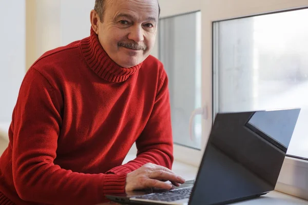 Um homem com a camisola vermelha a trabalhar no portátil junto à janela. Fazendo freelance na idade adulta . — Fotografia de Stock