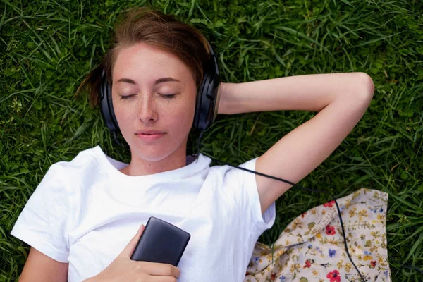 Una hermosa chica en auriculares se encuentra en la hierba verde en el parque y disfruta escuchando música con placer . — Foto de Stock