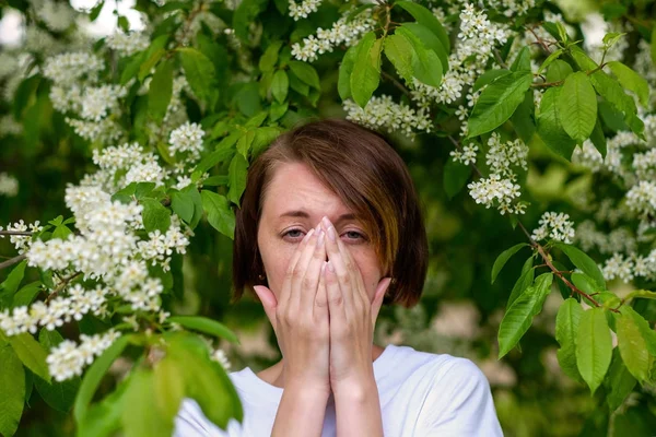 A beautiful girl stands by the blossoming bird cherry and sneeze — Stock Photo, Image
