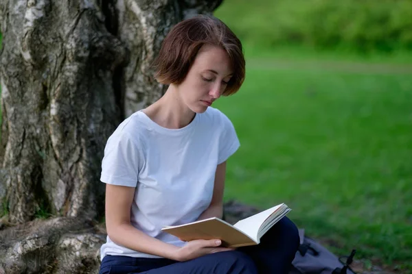 Una chica lee un libro en un parque sentado junto a un árbol sobre hierba verde . —  Fotos de Stock