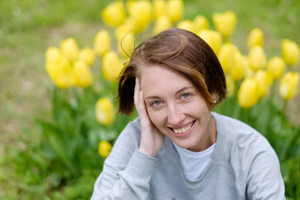 Uma menina com sardas senta-se na grama ao lado das tulipas amarelas — Fotografia de Stock