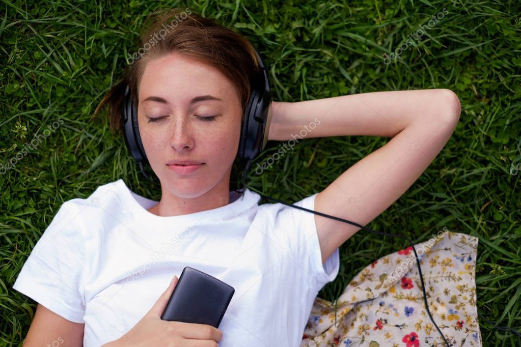 A beautiful girl in headphones lies on the green grass in the park and enjoys listening to music with pleasure.