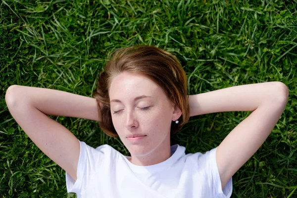 A beautiful girl with freckles lies on the green grass with her eyes closed and rests. — Stock Photo, Image