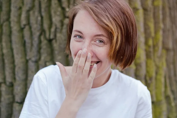 Lachende vrouw in het witte shirt met de rode haren over boom. — Stockfoto