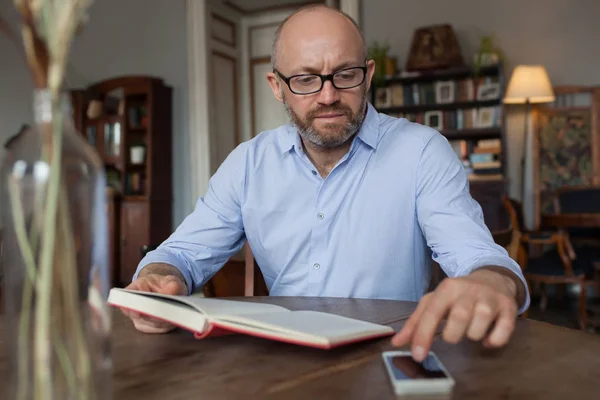 Ο άνθρωπος ανάγνωση βιβλίο εσωτερική και χρησιμοποιώντας το τηλέφωνό — Φωτογραφία Αρχείου