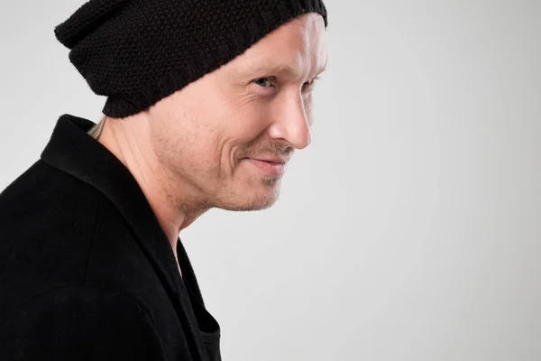 Jeune homme souriant en vêtements noirs debout et chapeau sur fond blanc regardant de côté — Photo