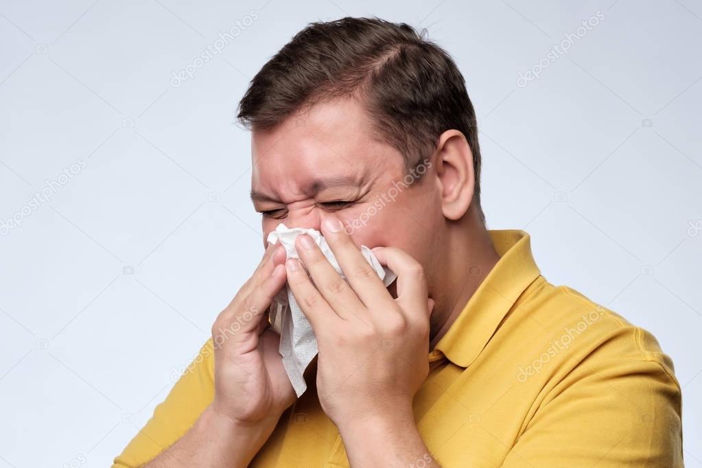 Mature fat caucasian man is ill from colds or pneumonia, sneezing in napkin.