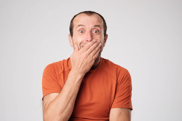 Mature caucasian man in orange t-shirt closing a mouth with hands on a light background.