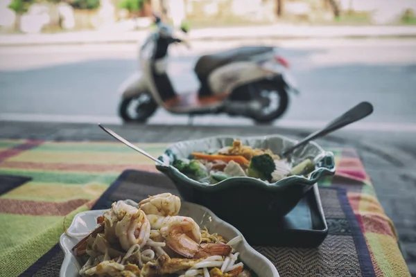 Two plates of spicy Thai food on the veranda on the street. In the background motorcycle. Stock Image