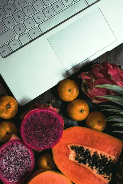 Тропические фрукты рядом с современным ноутбуком на деревянном фоне. Веганский образ жизни . — стоковое фото
