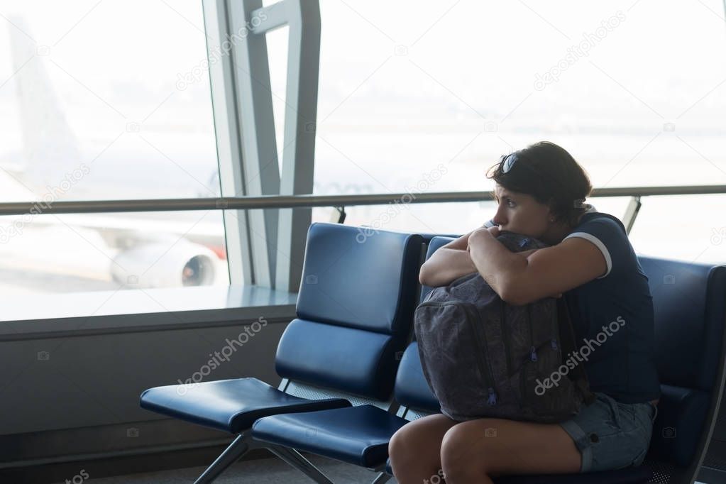 Tired caucasian woman in airport hall. Girl waiting her flight at airport terminal, holding her bag in hands.