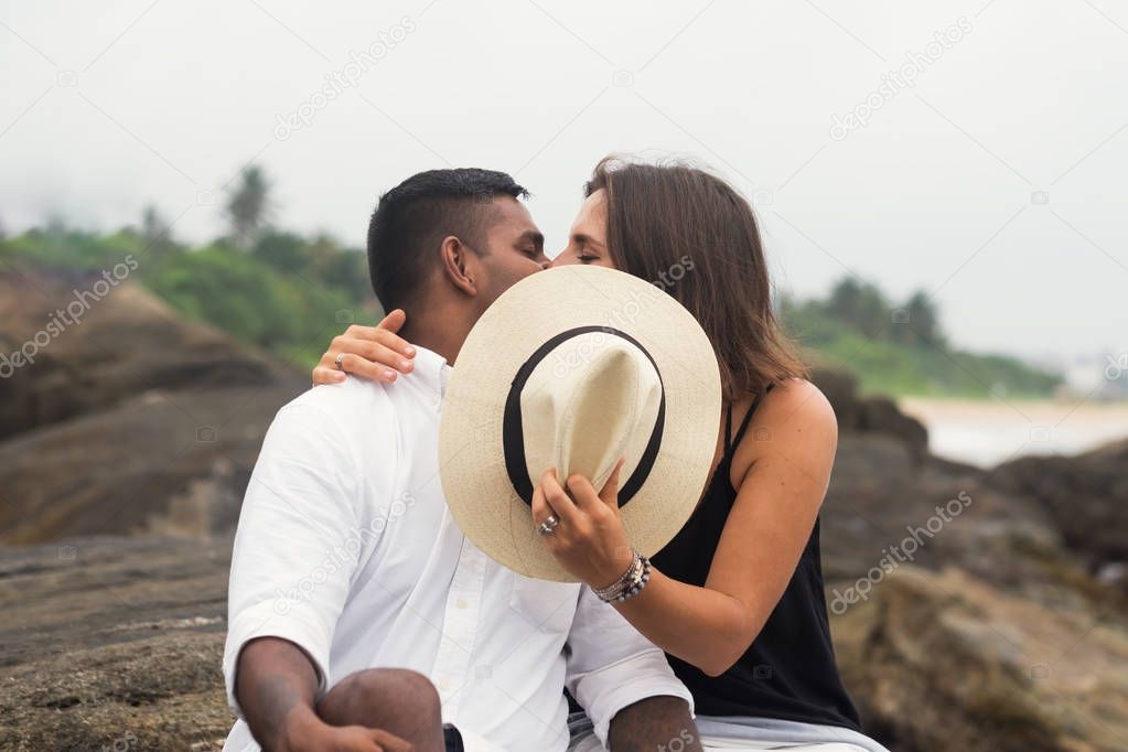 Young mixed race couple kissing sitting on stone. They close faces with hat.