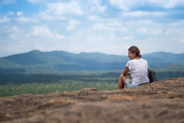 Junge Frau mit Rucksack sitzt auf einem Berg und blickt in einen Himmel mit Wolken — Stockfoto