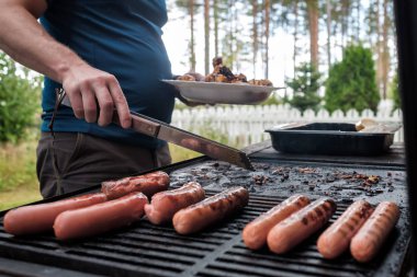 Genç beyaz adam elleriyle ızgarada sosis hazırlıyor.
